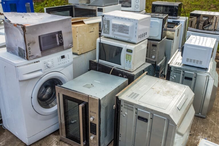 Large pile of old household appliances including washing machines, microwaves, ovens, and air conditioning units stacked outdoors.