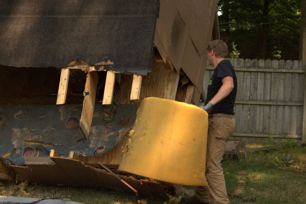 Ohio Junk Force worker carrying a large piece of yellow foam insulation while dismantling a damaged above-ground swimming pool structure in a backyard, with torn pool liner and wooden framing visible.