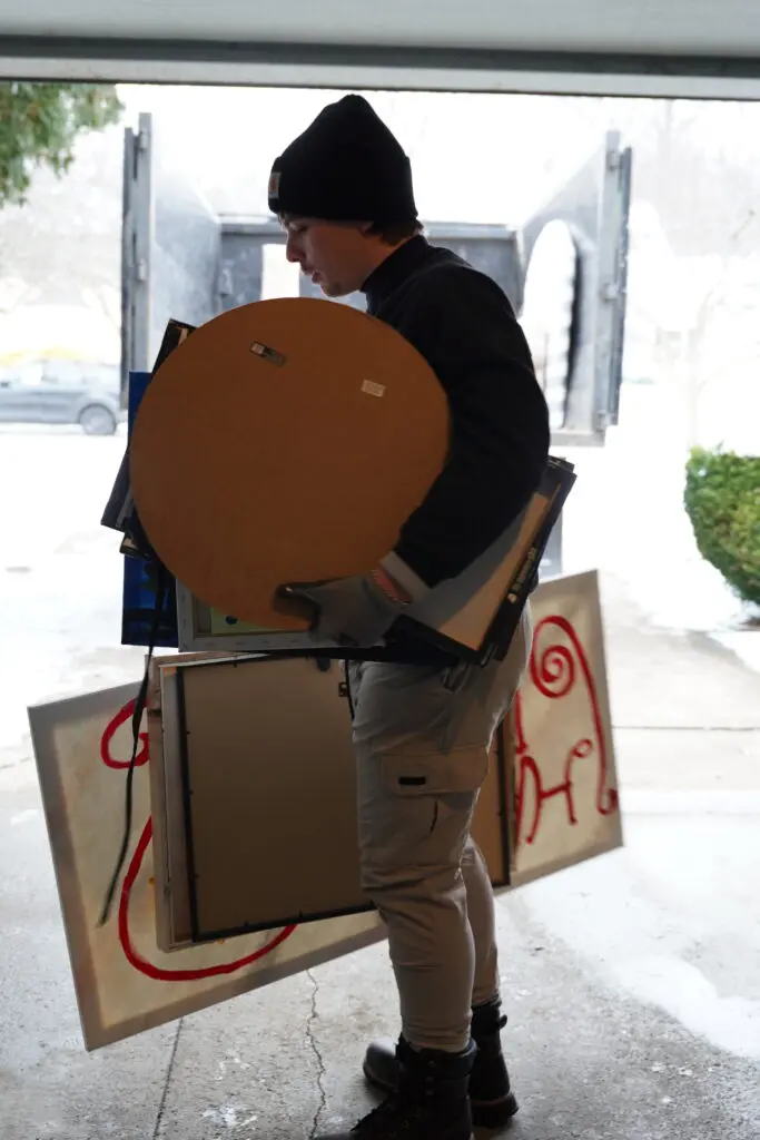 Worker in a garage carries a round tabletop, framed art, and other household items toward an open junk removal truck parked outside.