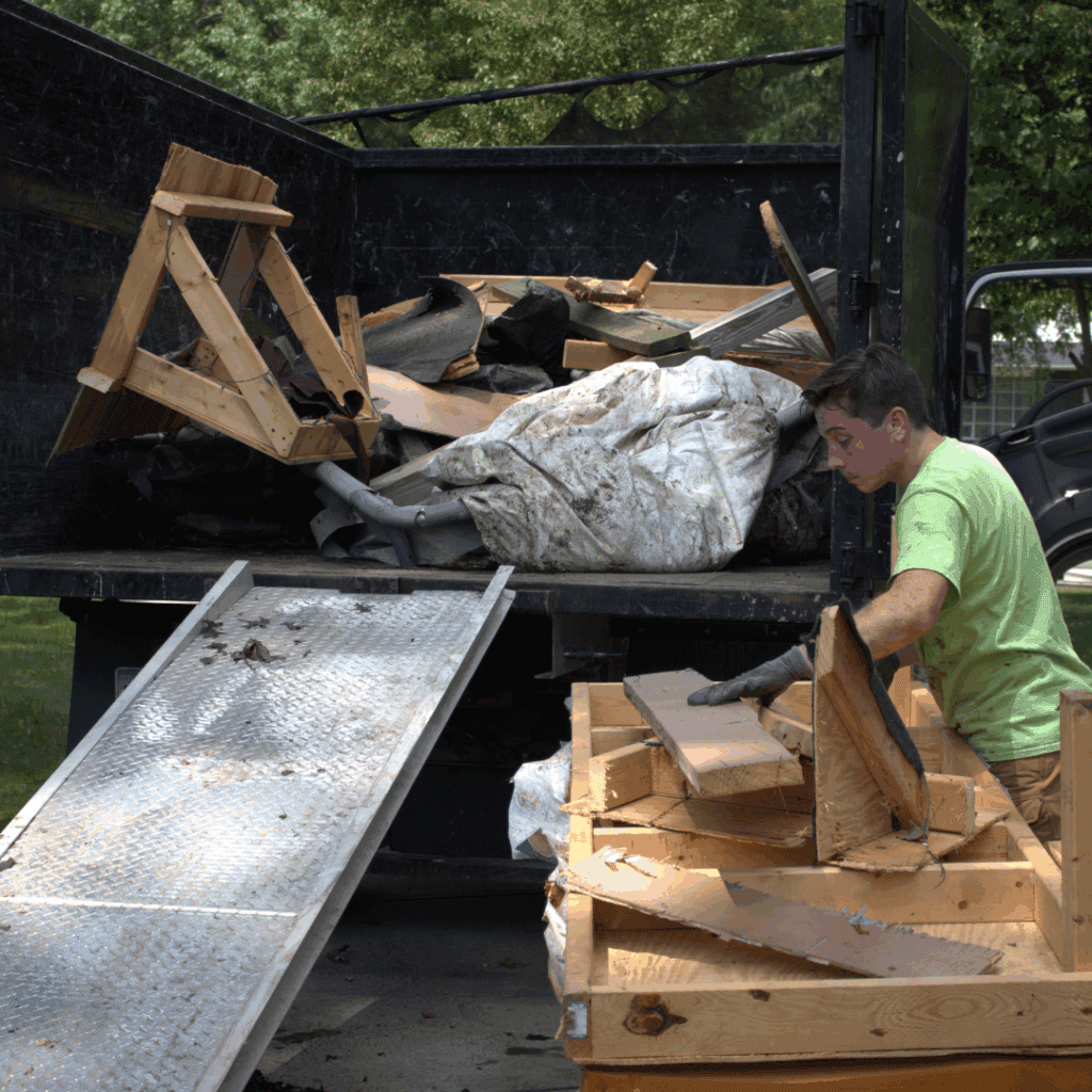 Worker in a green shirt loads broken wooden framing and construction debris into a black dump truck beside a metal loading ramp.