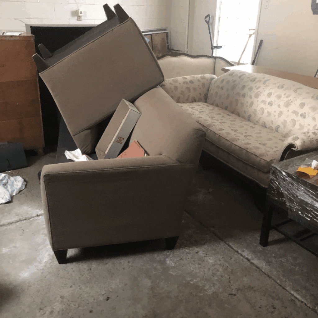 A chair, couch, cushions, and other furniture are stacked in a garage or storage area for removal.