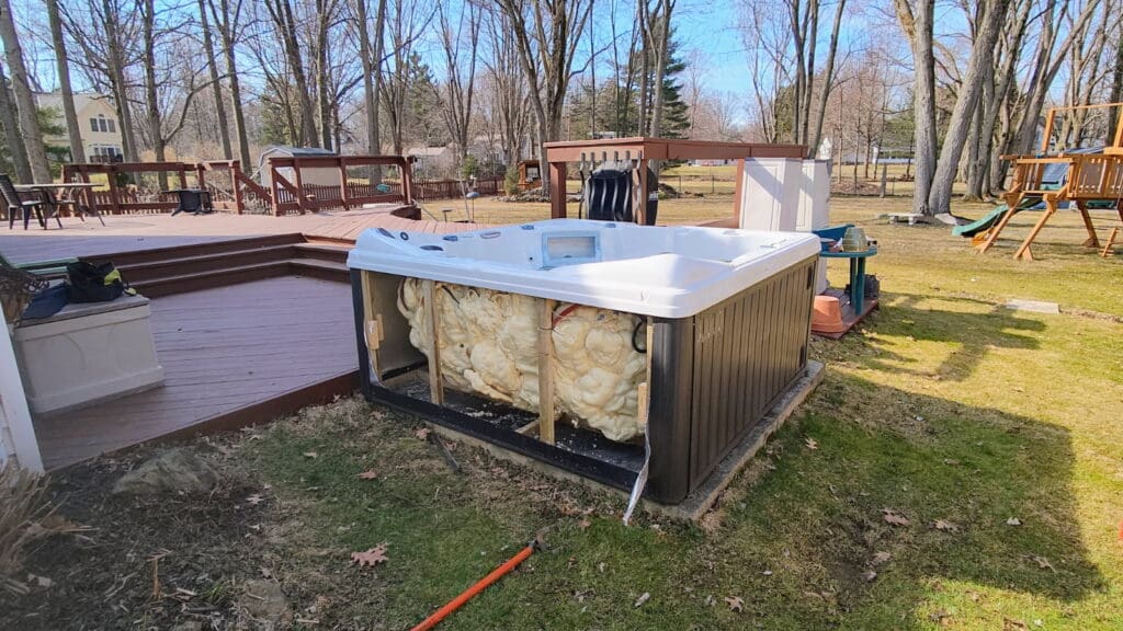 Outdoor hot tub with one side panel removed, exposing foam insulation and internal components, sits beside a raised deck in a backyard with tall leafless trees and a wooden playset.