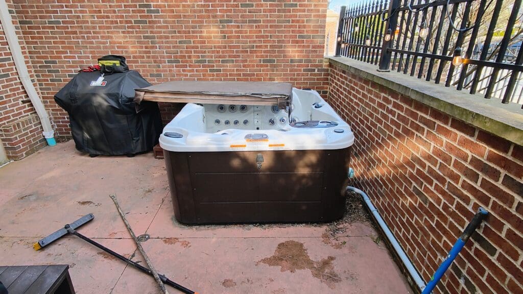 Outdoor hot tub with partially folded cover on a concrete patio between red brick walls, beside a covered grill, string lights, and long-handled cleaning tools.