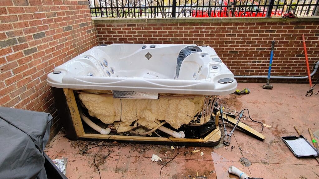 Large white hot tub with removed side panel exposing foam insulation, pipes, and wiring on a concrete patio enclosed by red brick walls, with tools scattered nearby.
