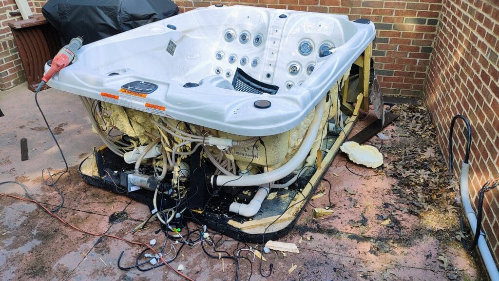Partially dismantled white hot tub with exposed foam insulation, plumbing, and tangled wiring on a concrete patio between brick walls, with a power tool resting on the rim.