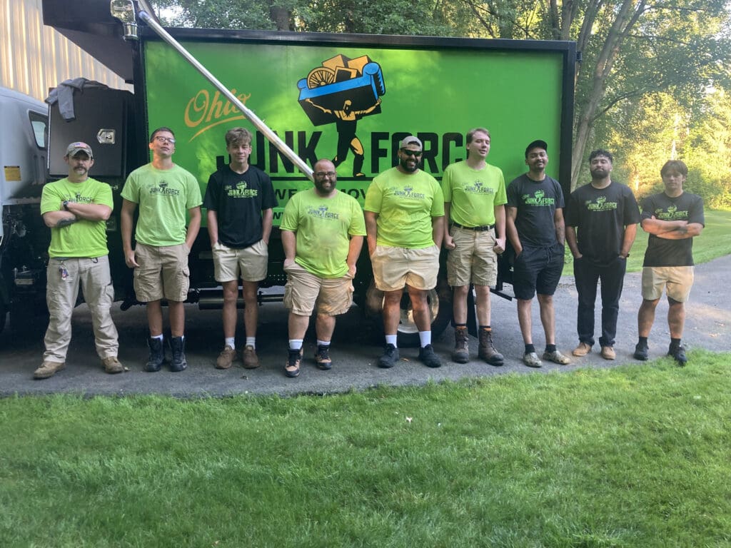 Group of nibe Ohio Junk Force crew members in green and black branded shirts stand smiling in front of bright green junk removal truck on driveway bordered by grass and trees.