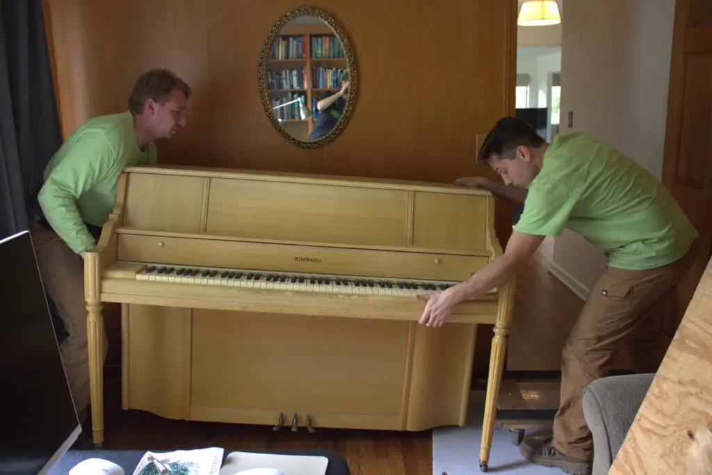 Two Ohio Junk Force workers in green shirts lift antique upright wooden piano indoors near oval mirror with bookshelves, lamp, and plywood protection on hardwood floor.