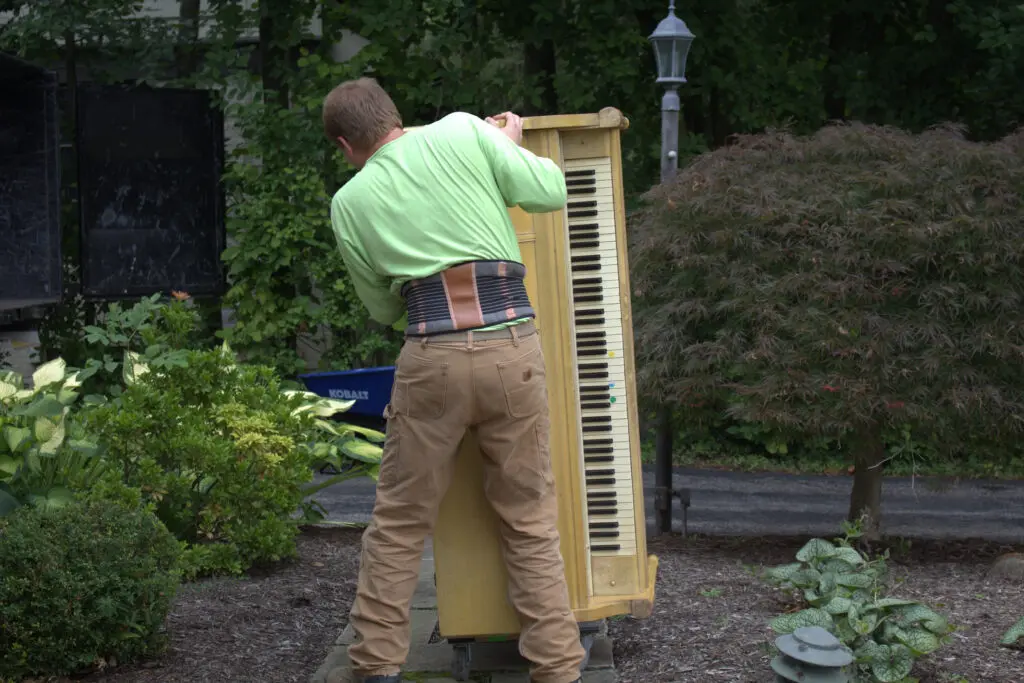 Ohio Junk Force worker in green shirt and back brace maneuvers upright wooden piano on dolly outdoors near shrubs, lamp post, and blue bin on mulch landscaping.