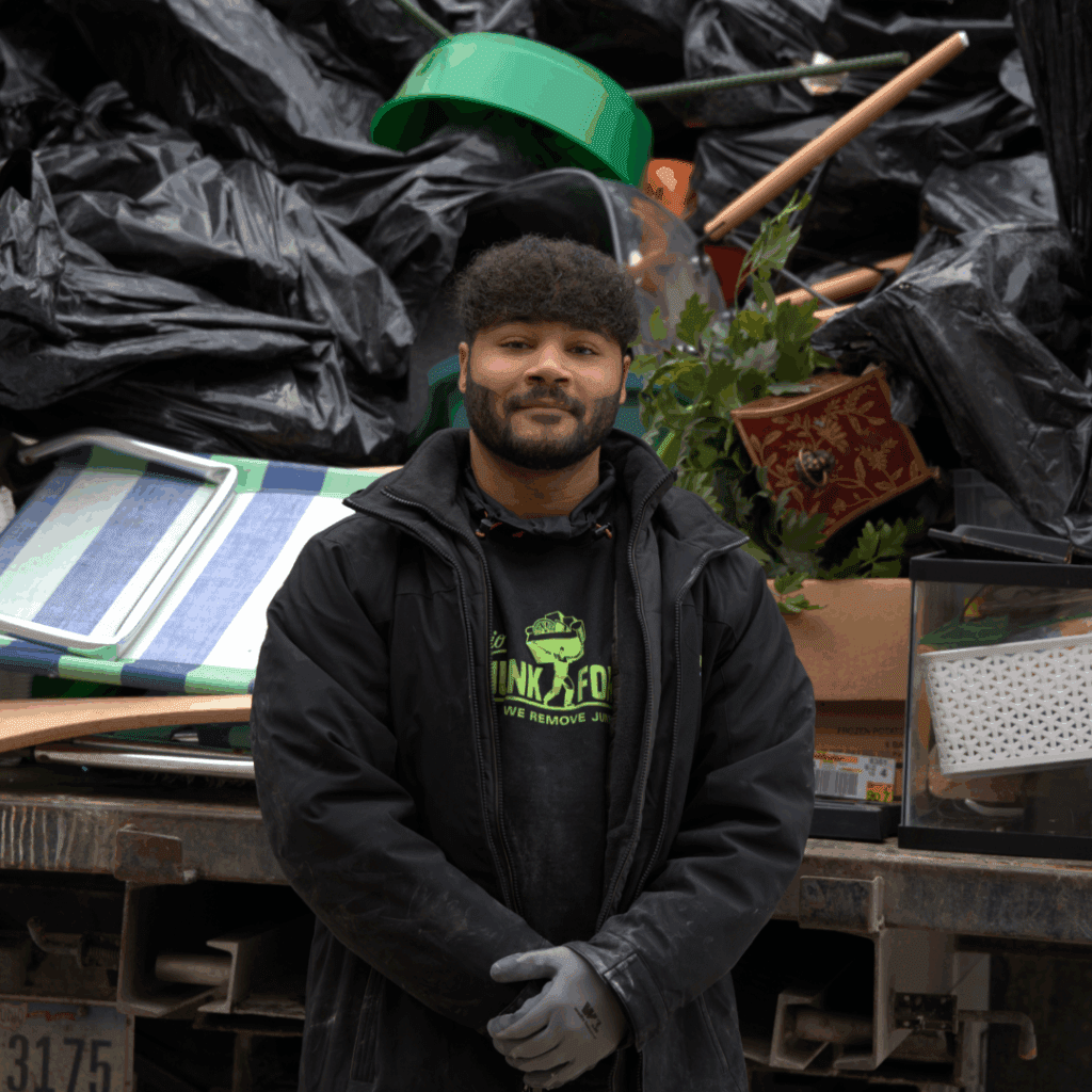 Junk removal worker in black jacket and gloves stands in front of a truck piled with black trash bags, outdoor furniture, and miscellaneous household debris.