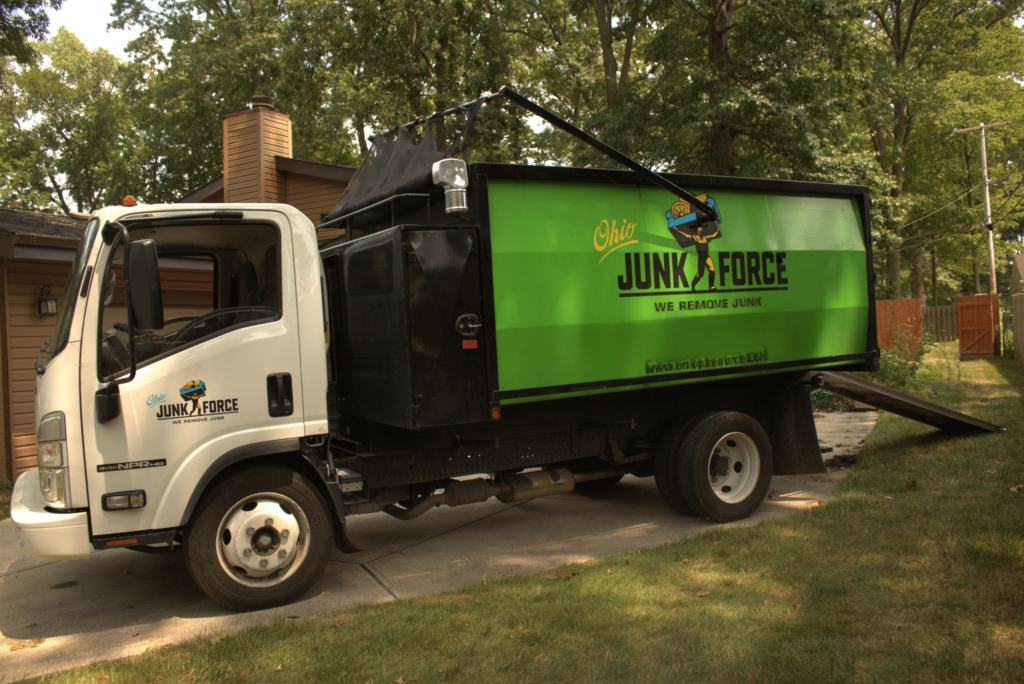 White Ohio Junk Force truck with green dump box and crane arm parked on driveway near house, featuring branded logo on side amid trees and fence.