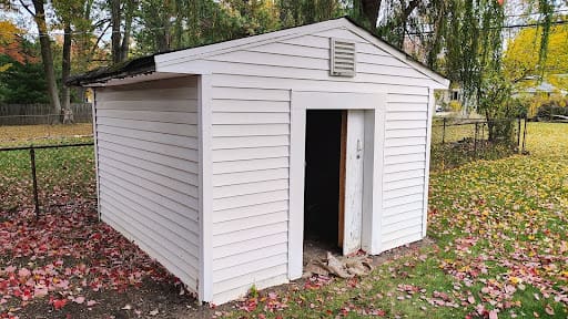 A small white storage shed with an open door standing in a backyard covered with fallen autumn leaves.