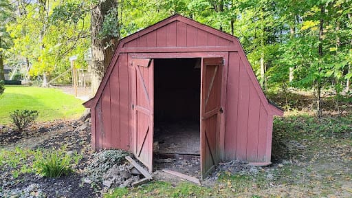 A weathered, red wooden storage shed with open double doors sitting in a grassy backyard surrounded by trees.