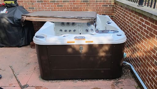 An empty, brown and white hot tub with a partially folded cover sitting on an outdoor patio next to a brick wall.