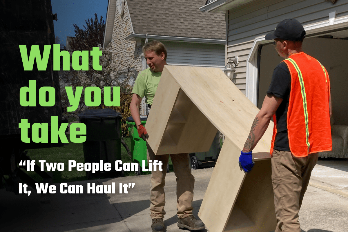 Two Ohio Junk Force workers—one in green shirt, the other in black shirt with orange vest—carry a tall wooden cabinet from house garage; green text reads "What do you take 'If two people can lift it. We can haul it.'"