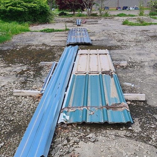 Stacks of blue and beige corrugated metal roofing panels lie on a cracked concrete lot surrounded by trees, shrubs, and a commercial building in the distance.