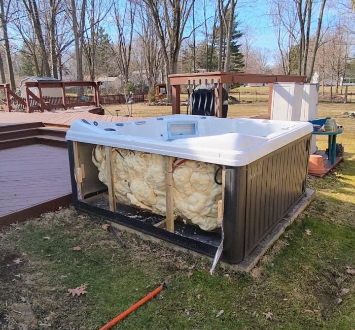 Outdoor hot tub with one side panel removed, exposing foam insulation and internal components, sits beside a raised deck in a backyard with tall leafless trees and a wooden playset.