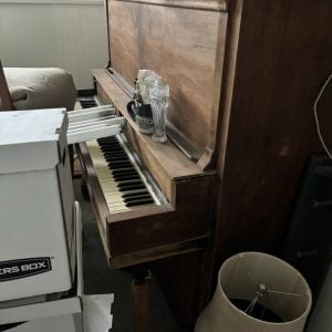 Vintage brown wooden upright piano in tight living room corner stacked with wicker baskets, CD cases, white U-Haul boxes, glass decanters, and fabric lamp shade nearby.