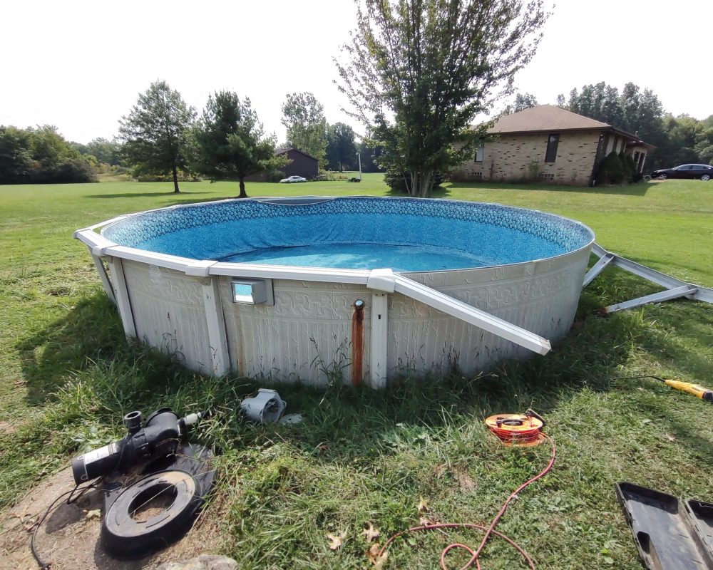 Above-ground swimming pool with blue water and patterned liner sitting on a grassy backyard, with pool equipment, hoses, and tools scattered on the ground around it.