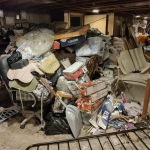 Heavily cluttered basement filled with piles of boxes, bags, furniture, an office chair, and various household items stacked throughout the space.