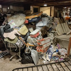 Heavily cluttered basement filled with piles of boxes, bags, furniture, an office chair, and various household items stacked throughout the space.