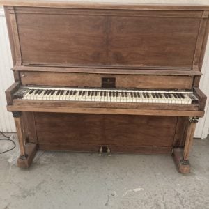 Vintage walnut upright piano with ivory keys and wooden pedals stands on concrete floor against beige wall, showing aged patina and classic cabinet design.
