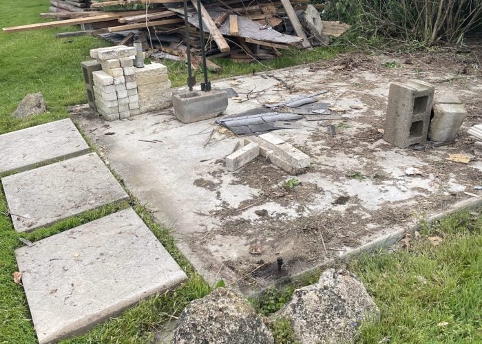 Backyard demolition site with a large pile of wooden debris, broken concrete blocks, paving stones, and construction waste scattered across the grass and concrete pad.