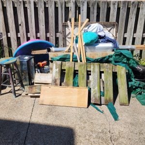 Pile of mixed outdoor items and scrap materials, including a wooden pallet, plastic storage bin, stool, cushions, and tarps, stacked on a concrete driveway beside a wooden fence.