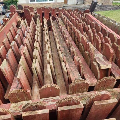 Large pile of dismantled red wooden fence sections and pickets stacked in a backyard, ready for removal.