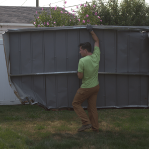 Worker in green shirt and brown pants carries a large gray metal shed wall panel across a grassy backyard near flowering bushes and utility lines.