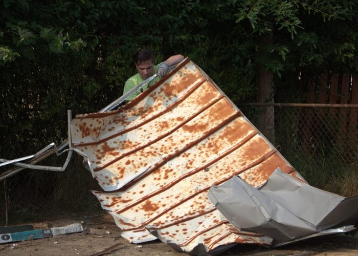 Ohio Junk Force worker in green shirt and gloves lifts large rusty corrugated metal sheet near chain-link fence and trees during junk removal job.