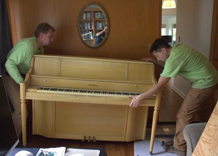 Two Ohio Junk Force workers in green shirts lift antique upright wooden piano indoors near oval mirror with bookshelves, lamp, and plywood protection on hardwood floor.