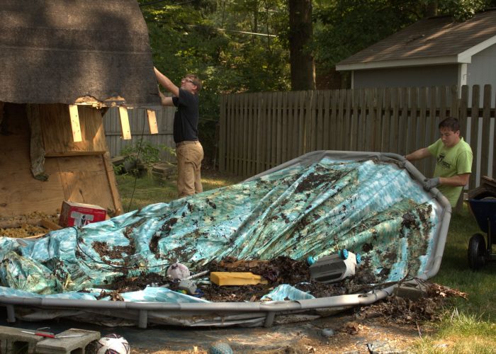 Two Ohio Junk Force workers dismantling and removing a large, dirty above-ground swimming pool from a backyard, with one man lifting the pool cover and the other handling the pool frame, surrounded by debris and tools.