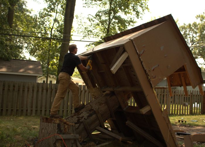 Ohio Junk Force worker standing on a tree stump while using a yellow power tool to dismantle a large, tilted wooden shed in a backyard, with the structure partially collapsed and debris scattered around.