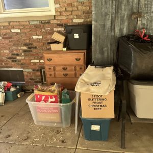 Cluttered garage corner with storage items including a wooden dresser, plastic bins, a cardboard box labeled “5 Foot Glittering Frost Christmas Tree”, and various household goods stacked against a brick wall.