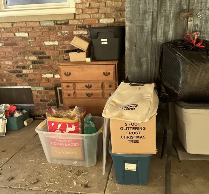 Cluttered garage corner with storage items including a wooden dresser, plastic bins, a cardboard box labeled “5 Foot Glittering Frost Christmas Tree”, and various household goods stacked against a brick wall.