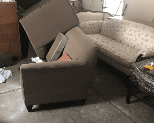 A chair, couch, cushions, and other furniture are stacked in a garage or storage area for removal.