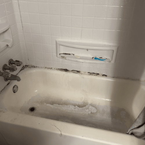 A dirty white bathtub and tiled wall with heavy black mold along the caulking, grime in the basin, and a razor in the soap dish.